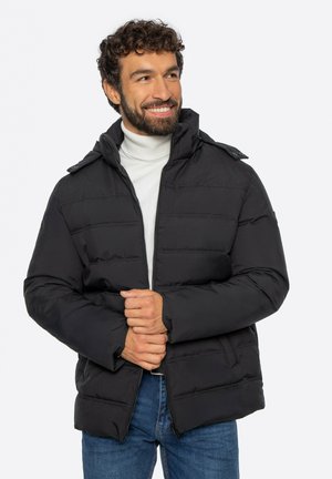 Uśmiechnięty mężczyzna z kręconymi włosami, ubrany w czarną kurtkę puchową na białym golfie i niebieskich dżinsach, trzymający kurtkę z przodu obiema rękami.
