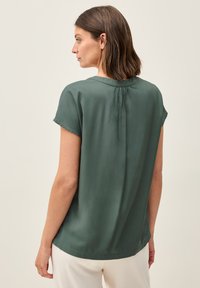 Blusa verde scuro a maniche corte con scollo rotondo, caratterizzata da un dettaglio plissettato sul retro e un tessuto leggero e morbido.