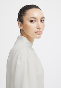 Blouse blanche à col montant avec des manches bouffantes, texture lisse et design minimaliste. Le modèle a une coiffure lissée et des boucles d'oreilles discrètes.