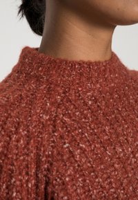 Ruosteenvärinen neulottu villapaita, jossa on korkea, resorimallinen kaulus. Pinta on pehmeä ja paksu, ja siinä on hienovarainen pilkullinen kuvio.