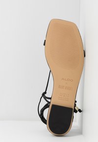 Vue inférieure d'une sandale noire à brides avec semelle beige montrant la taille 7,5 US, 38 EU, et la marque "ALDO x Rocky Barnes."