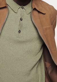 Zelené pletené polo s třemi tmavými knoflíky, pod které je oblečené světle hnědé semišové sako se zipem a boční kapsou.