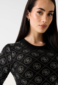 Kobieta o długich ciemnych włosach, ubraną w czarny top z drobnym białym wzorem kwiatowym w kształcie kółek oraz z pojedynczym wiszącym kolczykiem.