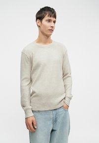 Camisa de manga longa em tom bege claro, texturizada, com um decote redondo, combinada com calças de ganga em azul claro. Cores neutras, ajuste casual, design minimalista.