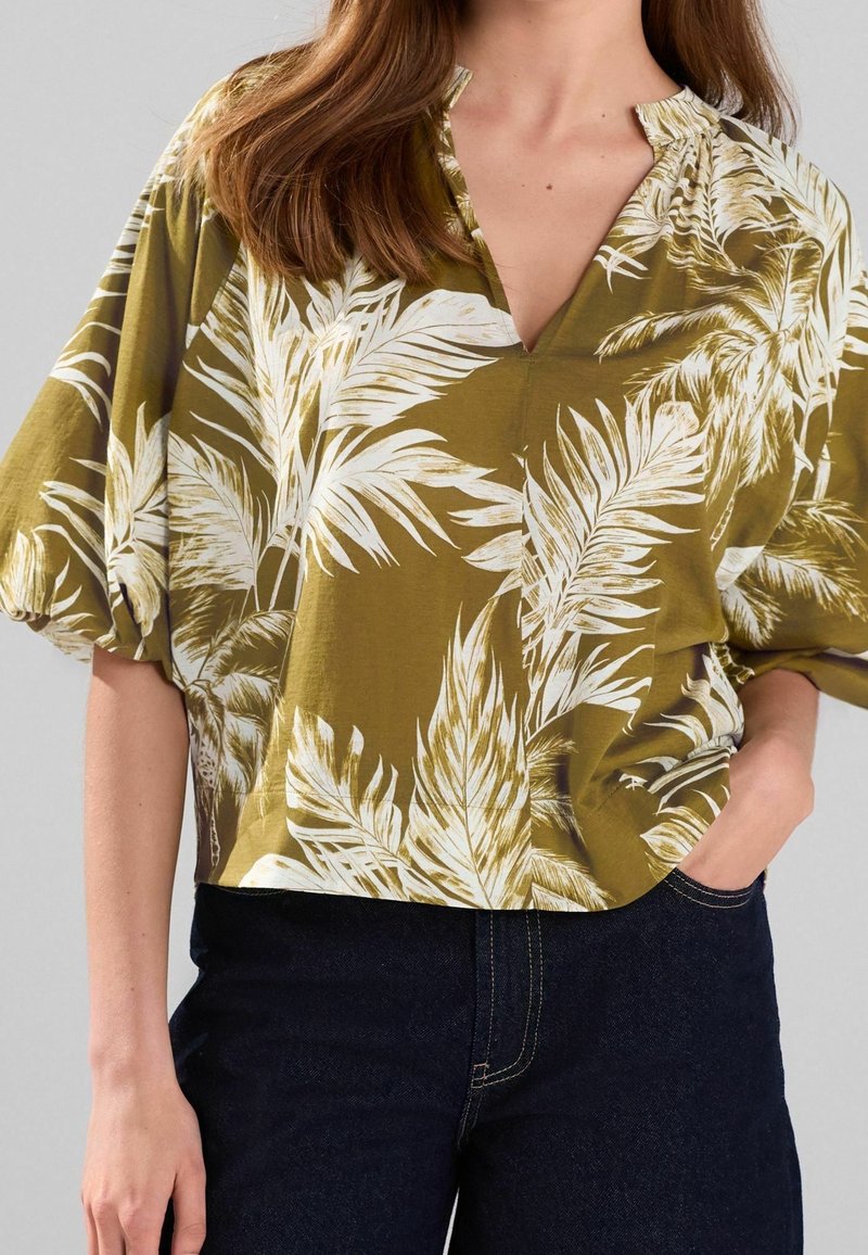 Blouse à manches courtes vert olive avec imprimé de feuilles de palmier blanches, décolleté en V et détails froncés aux épaules. Associée à un denim foncé.