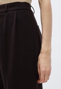 Pantalon noir sur mesure avec plis sur le devant, poches latérales et un logo brodé subtil sur le côté gauche. Texture de tissu lisse.