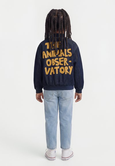 Παιδί με μακριά πλεξούδα που φοράει μπλε σκούρο μπουφάν με τη φράση "THE ANIMALS OBSERVATORY" σε κίτρινο, ανοιχτό μπλε τζιν και λευκά αθλητικά, στέκεται γυρισμένο προς τα πίσω.