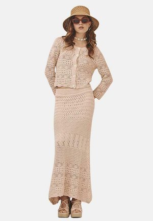 Conjunto de dos piezas de punto en beige claro, que incluye un cárdigan corto con botones y una falda larga con patrones intrincados; acompañado de un sombrero de paja.