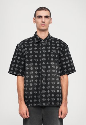 Chemise à manches courtes noire avec un motif en losanges gris, devant boutonné et poche poitrine unique, fabriquée en tissu denim avec une texture lisse.
