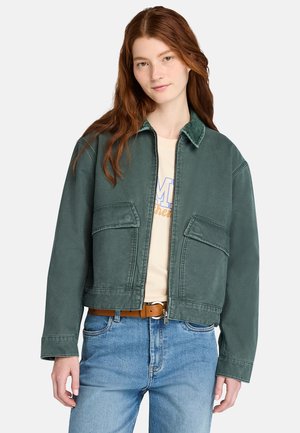 Groene cropped jas met een ritssluiting, twee voorkruisen en een kraag. Draagt over een lichtgekleurde blouse en blauwe jeans.