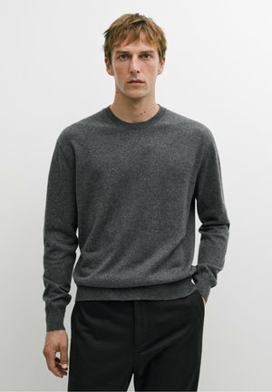 Giovane uomo con capelli castano chiaro che indossa un maglione semplice grigio scuro a maniche lunghe e pantaloni neri su uno sfondo chiaro uniforme.