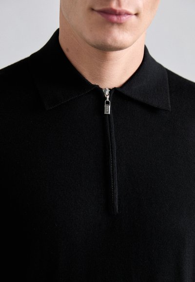 Sort zip-up polo sweater med spids krave. Lavet af blødt stof, med en metal lynlås og en struktureret design. Nærbillede af den øverste beklædningsgenstand.
