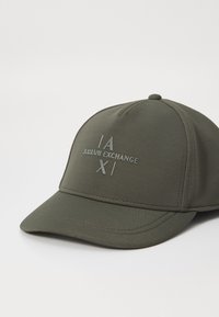 Olivengrønn baseballcap laget av teksturert stoff, med en hevet logo "ARMANI EXCHANGE" og kryssete linjer på frontpanelet.