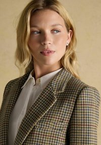 Béžový blazer s kožuchovým vzorom a červenými a modrými akcentmi, v kombinácii s bielou košeľou s golierom. Má štruktúrované ramená a textúrovaný vzor.