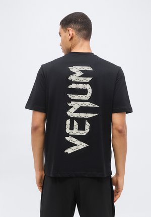 Άνδρας με κοντά μαλλιά φοράει μαύρο μπλουζάκι με μεγάλο κατακόρυφο λευκό κείμενο "VEJA" στην πλάτη, πάνω σε απλό φόντο.