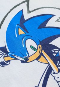 T-shirt blanc avec un graphique bleu représentant un personnage aux yeux verts et aux accents jaunes. Le design comprend des contours nets et des couleurs vives.