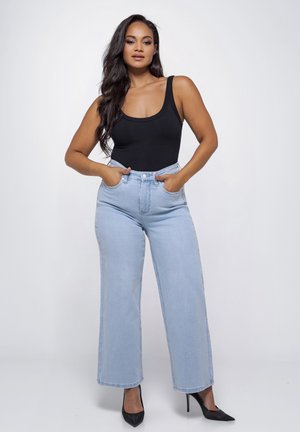 Femme debout les mains dans les poches d'un jean large taille haute bleu clair, portant un haut noir sans manches et des talons hauts noirs.