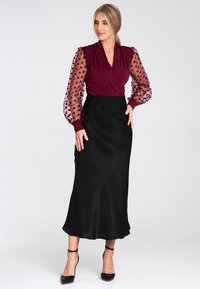 Bordowa sukienka z przezroczystymi, kropkowanymi bufkami, dekoltem w stylu opaskowym oraz gładką czarną spódnicą, zakończona falującym dołem i eleganckimi czarnymi szpilkami.