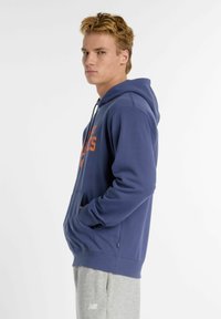 Felpa con cappuccio blu navy con grafica arancione, cappuccio con cordino, tasca a marsupio frontale e polsini e orlo a costine. Abbinata a pantaloni della tuta grigi.