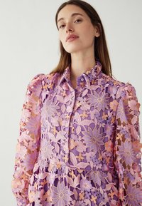 Violet siuvinėta bliuska su gėlių nėriniais ir rožinėmis detalėmis, mygtukų uždarymu ir struktūrizuotu apykakle. Permatomi ilgi rankoviai.