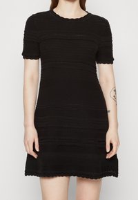 Abito in maglia nera con maniche corte, caratterizzato da una texture a coste e bordi ondulati. Il motivo include righe orizzontali e un colletto rotondo.