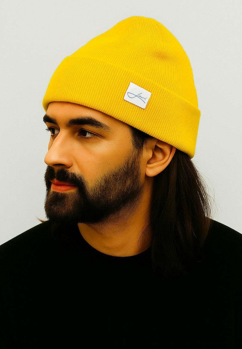 Yellow ribbed beanie with a folded cuff featuring a small white label. Worn by a person with a beard and long hair against a neutral background.
