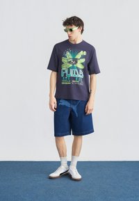 Νεαρός άνδρας φορώντας μπλε μπλούζα με γραφικό λογότυπο Puma, τζιν σορτς με μεγάλο λογότυπο Puma, λευκά αθλητικά παπούτσια, λευκές κάλτσες και νέον πράσινα γυαλιά ηλίου, στέκεται σε μπλε πάτωμα.