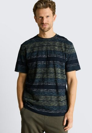 Mężczyzna o jasnobrązowych włosach ubrany w granatowo-oliwkową koszulę w paski z krótkim rękawem, stoi z rękami w kieszeniach.