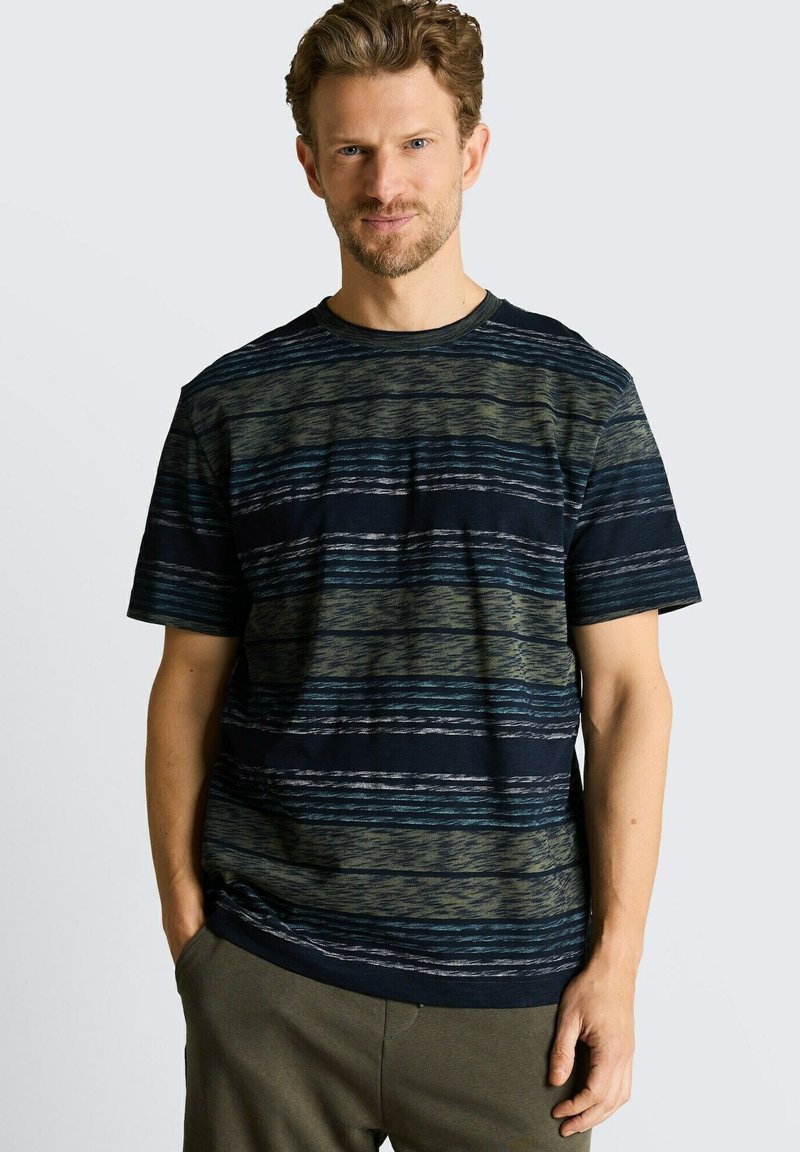 Hombre con cabello castaño claro que lleva una camisa de manga corta a rayas azul marino y verde oliva, de pie con las manos en los bolsillos.