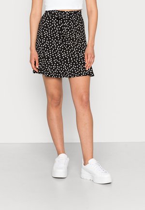Kvinde iført en sort nederdel med hvide polkaprikker og en sløjfe i taljen, kombineret med hvide platformsneakers og en hvid cropped top.