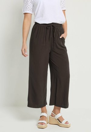 Mujer con pantalones negros holgados de corte corto con cintura ajustable, blusa blanca de manga corta con ojales y sandalias de cuña blancas.