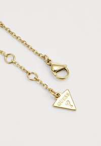 Brățară din aur cu închizătoare tip homar și un charm triangular gravat cu "GUESS" și un semn de întrebare. Textură netedă, design elegant.