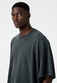 Tumši pelēks, pārāk liels T-krekls ar mīkstu, teksturētu audumu, klasisku apaļu kakla izgriezumu un īsām piedurknēm. Minimalistisks dizains bez rakstiem vai akcentiem.