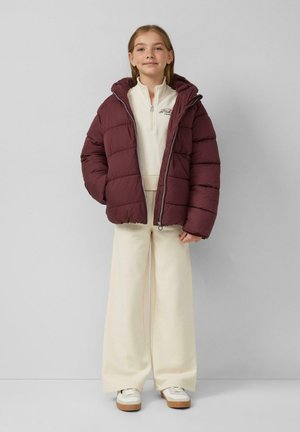 Bordeaux pufferjack met een capuchon, voorzien van een ritssluiting en een gekruist patroon, gedragen over een crèmekleurige coltrui en een wijde broek.