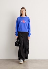 Felpa blu con logo rosso 'Calvin Klein', pantaloni cargo neri, sneakers argento e una piccola borsa nera. Tessuto liscio e vestibilità rilassata.