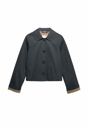 Mujer rubia vista desde atrás, vistiendo una chaqueta negra con puños a cuadros y pantalones beige sobre un fondo liso.