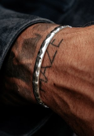 Silver metal bracelet with a hammered texture, worn around a wrist with visible tattoo, complementing a dark fabric background.