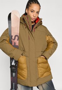 Brun pufferjakke med polstrede guld-gule detaljer, store lommer og høj krave. Modellen holder et ski på den ene side.