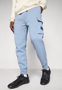 Person wearing light blue cargo sweatpants with side pockets and black sneakers, standing against a plain light background.