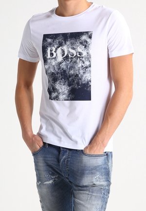 Homme portant un T-shirt blanc avec un motif graphique abstrait noir et blanc et le texte « BOSS HUGO BOSS », associé à un jean bleu déchiré.