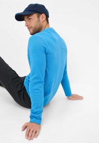 Blauw langshirt met ronde hals, gecombineerd met donkere broeken. De persoon draagt een marineblauwe pet en zit tegen een witte achtergrond.