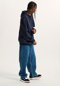 Felpa con cappuccio navy, vestibilità rilassata, abbinata a jeans oversize di un blu chiaro. Sneakers bianche e nere con suola in gomma. Stile streetwear casual.