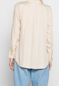Light beige long-sleeve shirt with a small repeating pattern, buttoned cuffs, and a curved hem, made from a soft, smooth fabric.