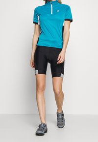 Tyrkysový cyklistický dres s krátkým rukávem a zipovým límcem, v kombinaci s černými cyklistickými šortkami s bílými pruhy a vycpaným sedákem.