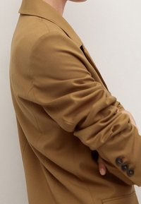 Blazer en tissu marron avec une coupe ajustée, un revers cranté et des détails de poignets boutonnés. La texture semble lisse et légèrement froissée.