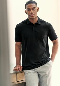 Camisa polo negra con cuello y mangas cortas, combinada con pantalones gris de cuadros, sobre un fondo claro.