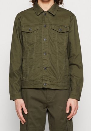 Homme portant une veste boutonnée vert olive avec poches à rabat sur la poitrine et un pantalon assorti, debout devant un fond uni.