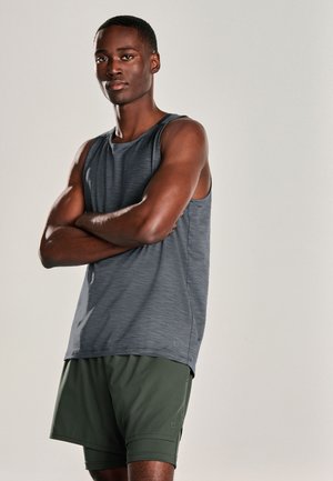 Hombre atlético con los brazos cruzados, vistiendo una camiseta sin mangas gris y pantalones cortos verde oscuro sobre un fondo liso.
