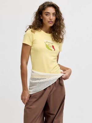 Femme aux cheveux bouclés portant un t-shirt jaune imprimé de piments et un pantalon marron, ajustant une ceinture ou un accessoire en maille blanche.