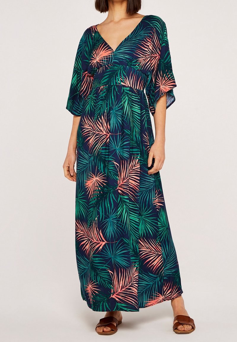 Femme portant une robe maxi bleu marine avec motif de feuilles de palmier vertes et corail, col en V, manches larges et sandales marron ouvertes à l'avant, debout sur un fond neutre.